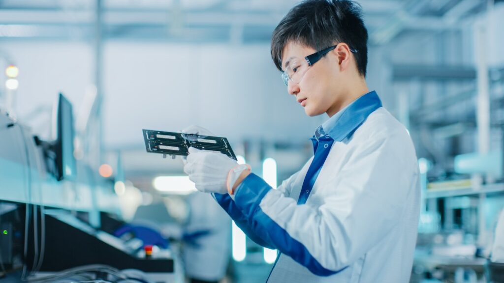 High-Tech Factory: Quality Control Engineer Checks Electronic Printed Circuit Board it for Damages. In the Background Assembly Line for PCB with Surface Mount Pick and Place Technology.